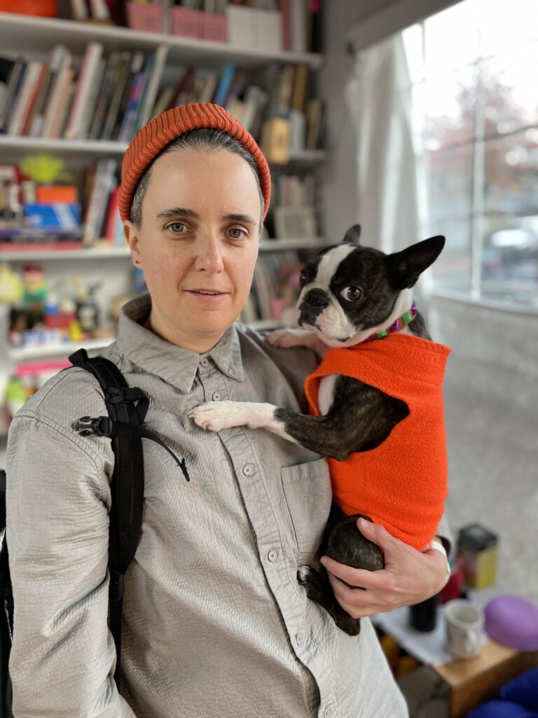 A portrait of Cait McKinney wearing a grey button-up shirt, orange wool beanie, holding a boston terrier who is wearing an orange fleece sweater. They are posted in front of a bookshelf, beside a large window.